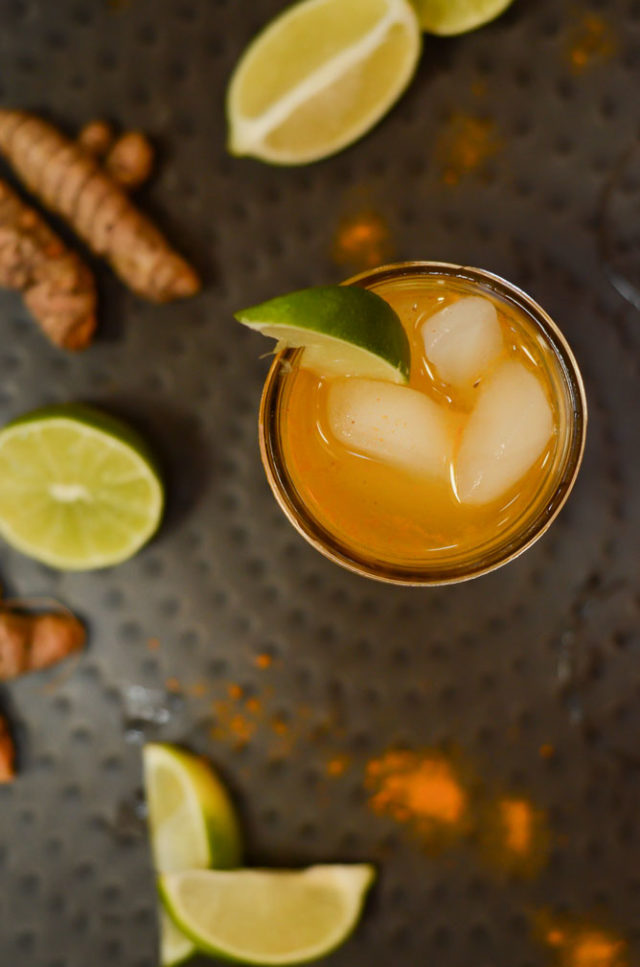 An overhead shot of a turmeric cocktail garnished with a lime and served on a black metal tray.