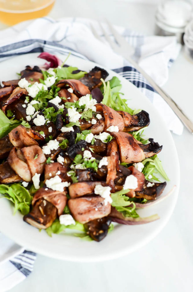 A large serving platter of Bacon-Wrapped Portobello Mushrooms with Goat Cheese over a bed of mixed greens.