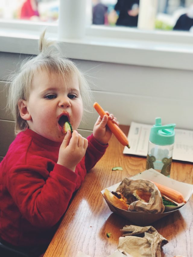 A toddler enjoying raw veggies sticks for dinner. Toddler meal planning is possible even when eating out!