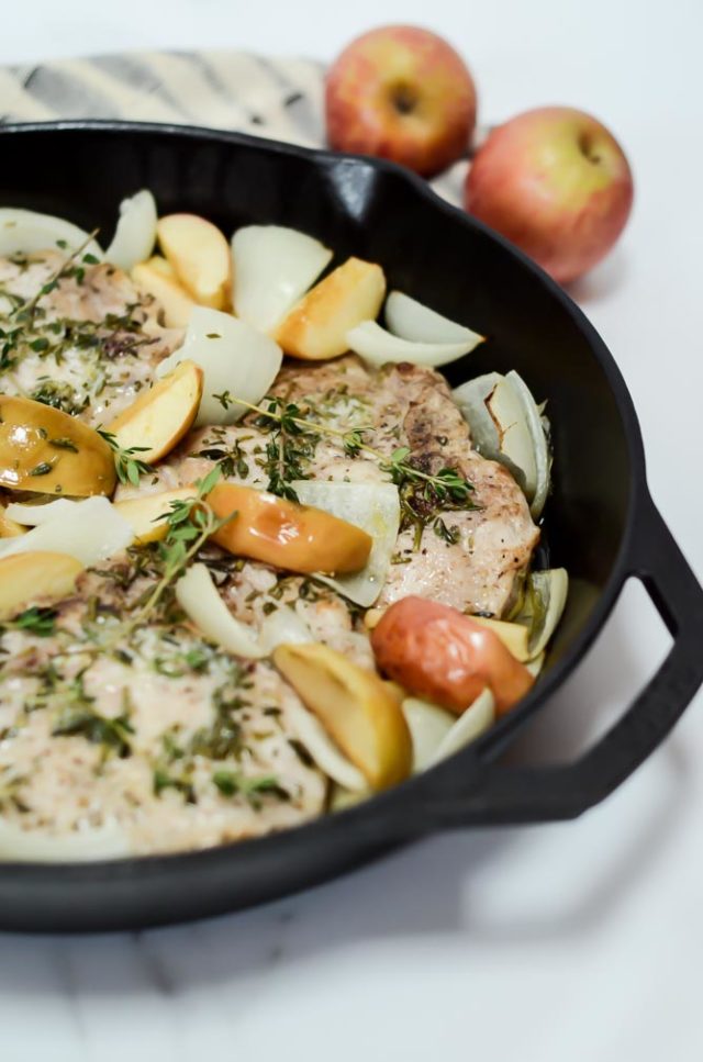 A close-up view of Apple and Herb Pork Chops in a cast iron skillet, with whole apples in the background.