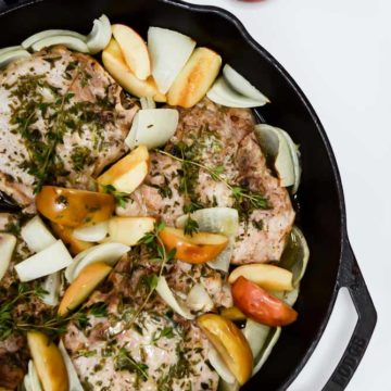 A beautiful skillet of Weeknight One-Pan Apple and Herb Pork Chops tied off with a black and white gingham hand towel, with a pile of fresh apples off to the side.
