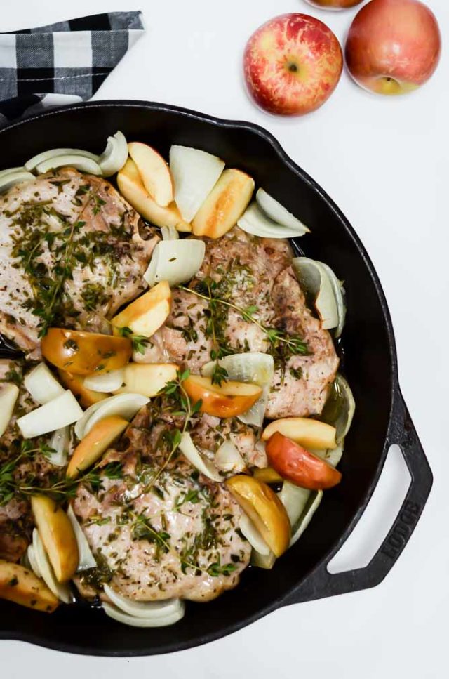 A beautiful skillet of Weeknight One-Pan Apple and Herb Pork Chops tied off with a black and white gingham hand towel, with a pile of fresh apples off to the side.