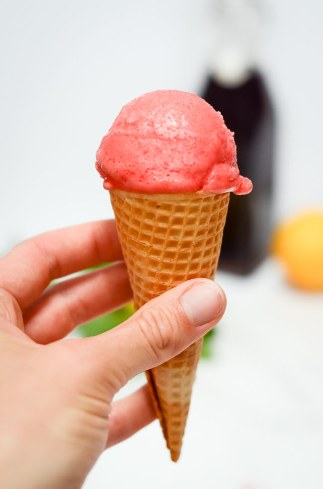 A hand holding a scoop of Raspberry Meyer Lemon Sorbet in a sugar cone.