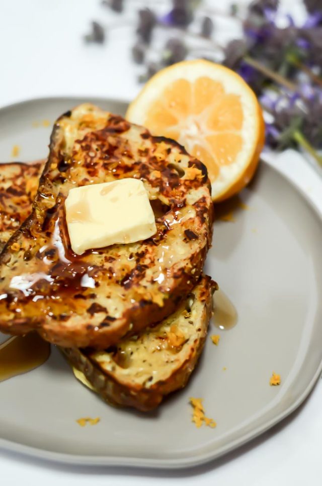 An overhead shot of two pieces of lemon ricotta French toast on a plate with a lemon wedge, topped with a pat of butter and syrup, flanked by purple flowers.