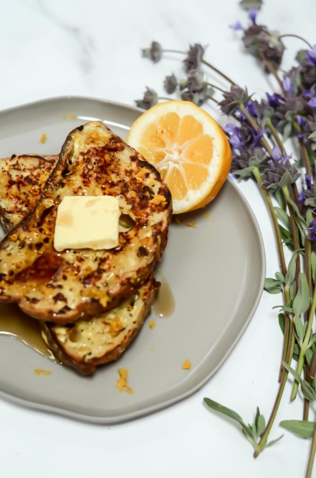 Another overhead shot of lemon ricotta french toast on a grey plate, covered with butter and syrup, with a lemon wedge for garnish.