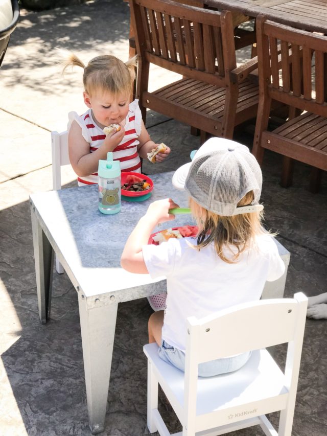 Two toddlers enjoying an outdoor lunch at a kids' table.