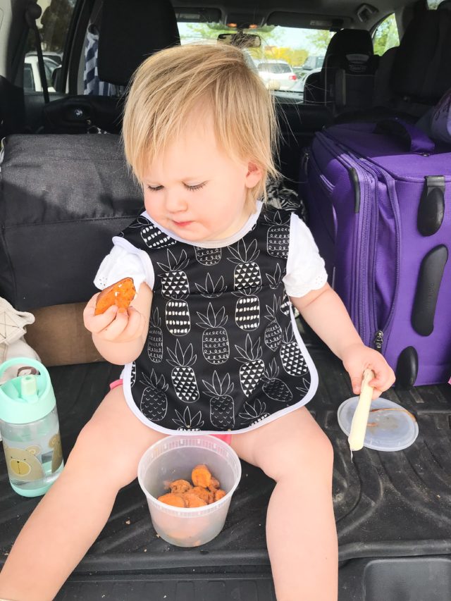 A toddler sitting in the back of a car eating some sweet potatoes.