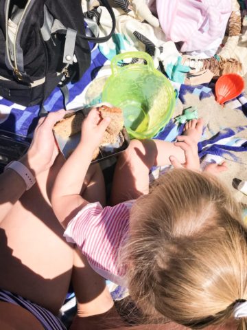 A toddler having a lunch picnic on the beach. Creative lunch ideas are important for exposing your child to a wide variety of foods!