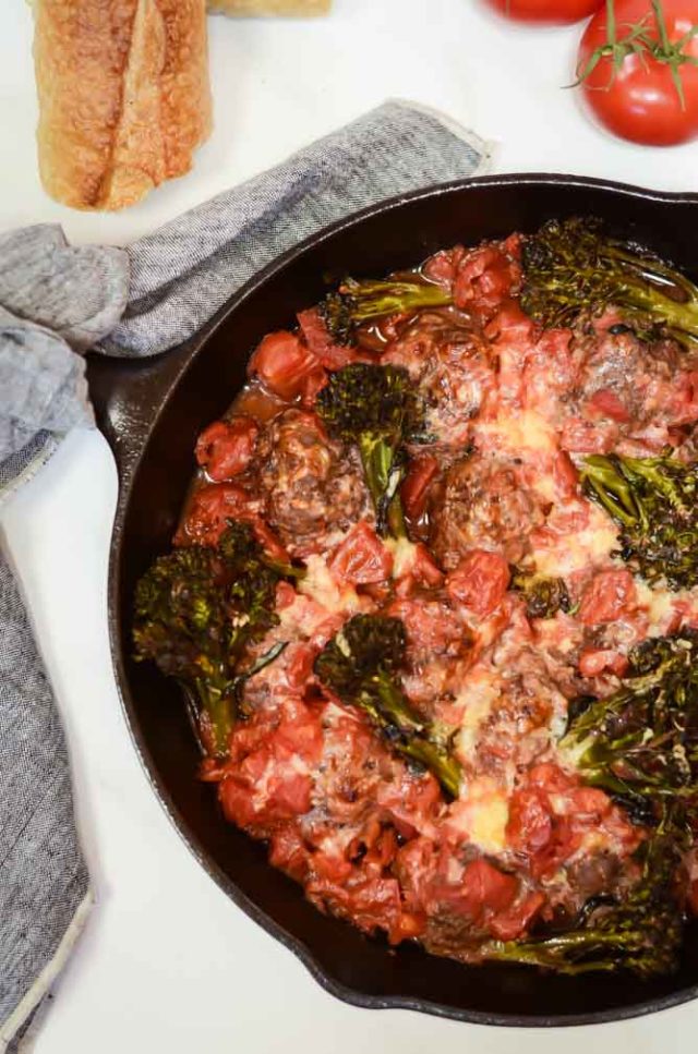An overhead shot of a pan of bison meatballs with broccolini and tomato sauce. The pan is tied off with a hand towel and there are tomatoes and bread on either side.