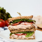 A Quick-and-Healthy Tuna Salad Sandwich cut in half on a plate with produce behind it.