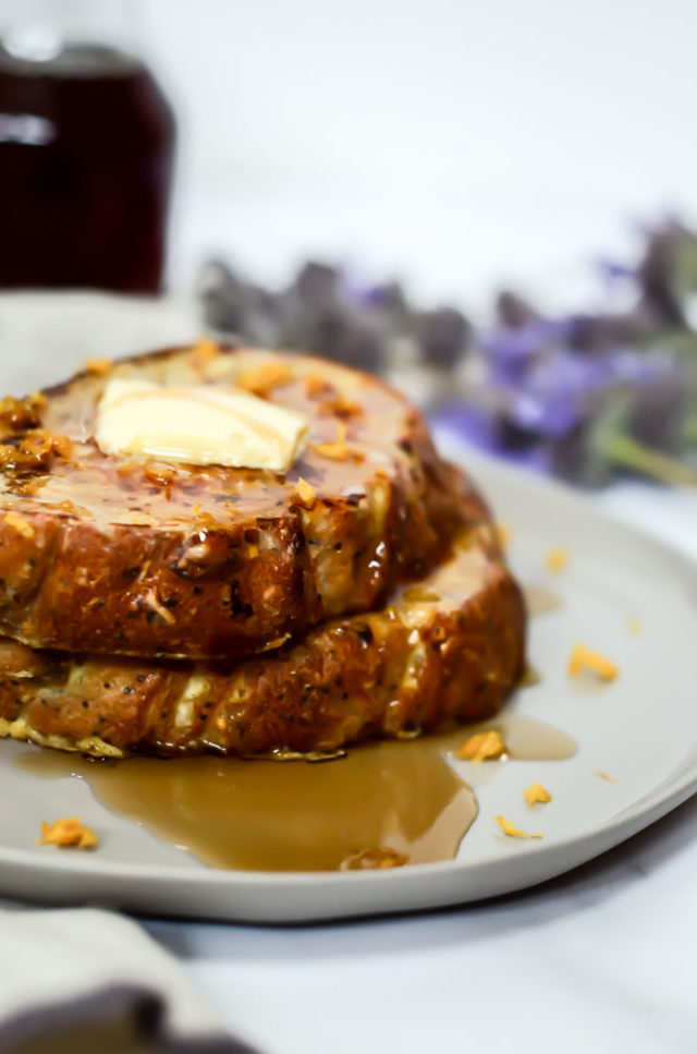 Two slices of lemon ricotta French toast are stacked on a plate, topped with a pat of butter and drizzled with syrup.
