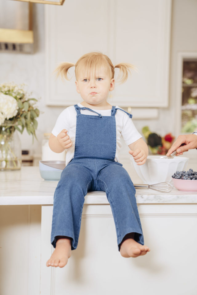A toddler sitting on a kitchen counter eating blueberries with a quizzical look on her face.