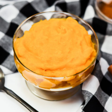 Homemade pumpkin puree in a glass dish surrounded by a black spoon, black and white checked napkin and a small bowl of ground cinnamon.