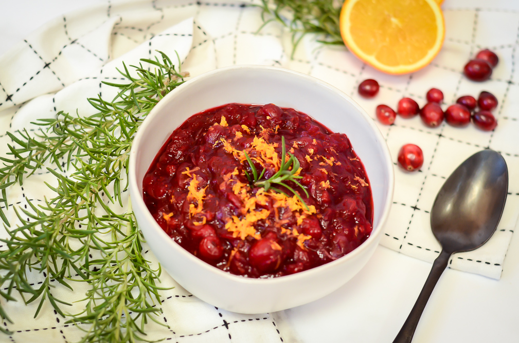 A bowl of cranberry sauce flanked by fresh rosemary, cranberries and orange.