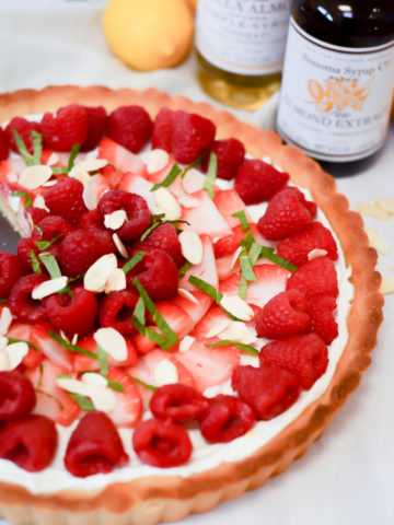 A Greek Yogurt Berry Tart with Almond Crust surrounded by bottles of almond extract and almond syrup, plus fresh lemons.