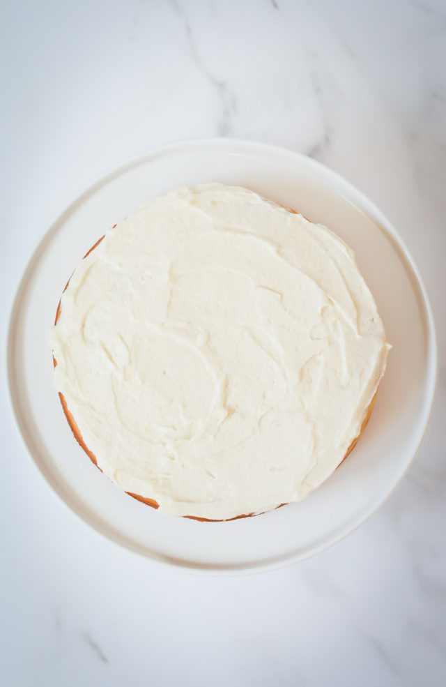 An overhead shot of the frosted bottom layer of a vanilla cake on a white cake stand.