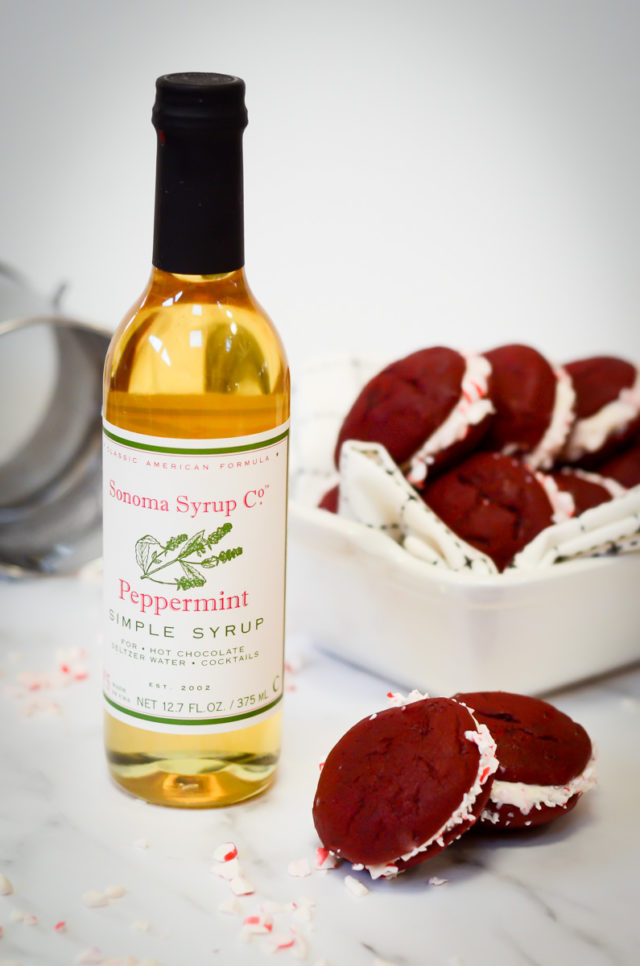 A batch of red velvet whoopie pies next to a bottle of peppermint syrup.