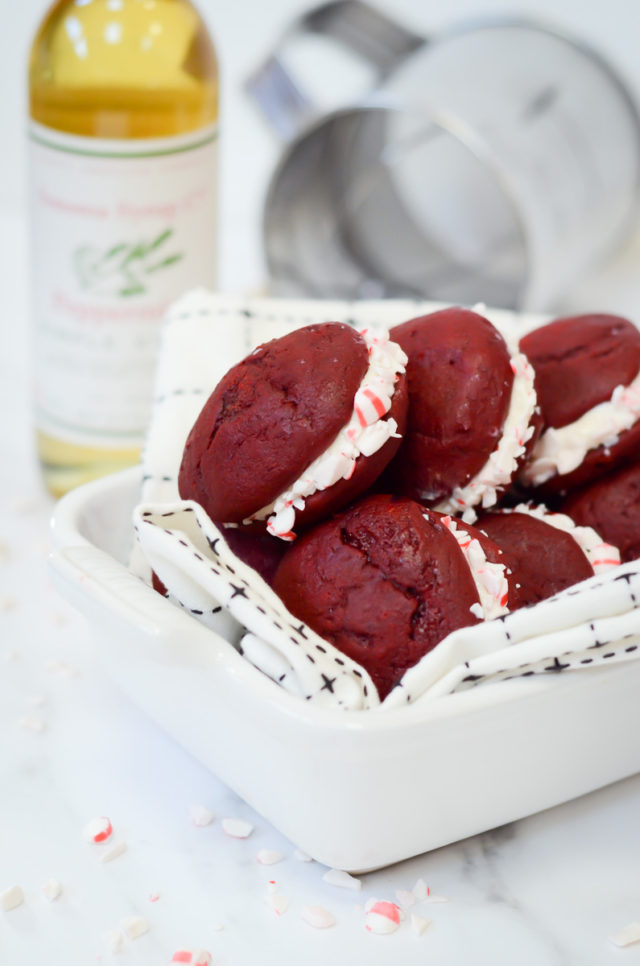 A dish full of peppermint whoopie pies with a bottle of peppermint syrup and a sifter in the background.