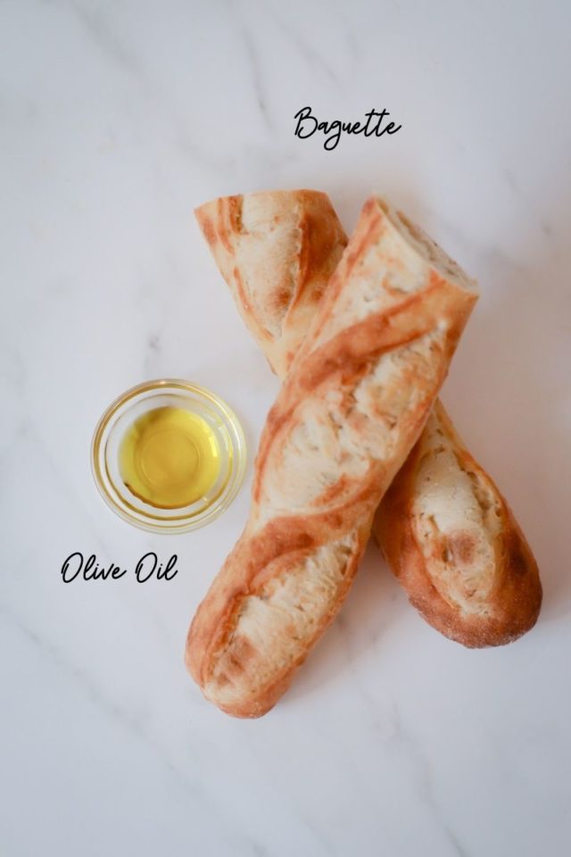 A baguette cut in half and a dish of olive oil on a marbled surface.