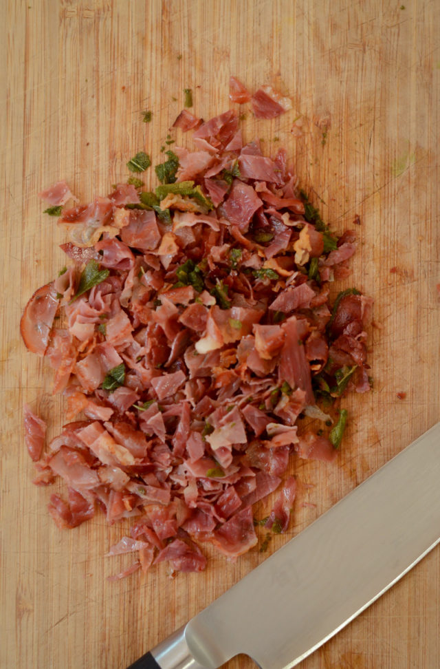 Chopped prosciutto and sage with a knife on a wooden cutting board.